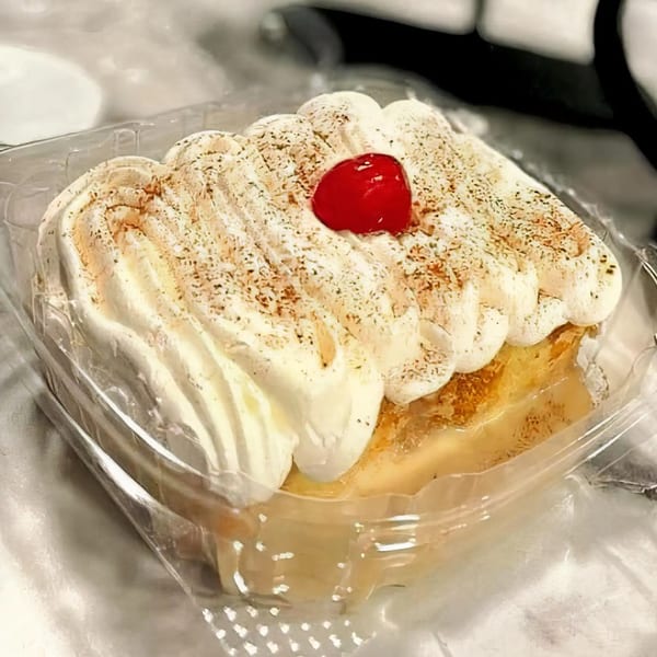 Moyitos Cafe & Grill's tres leches cake with whipped cream, cinnamon, and a cherry in a plastic container.